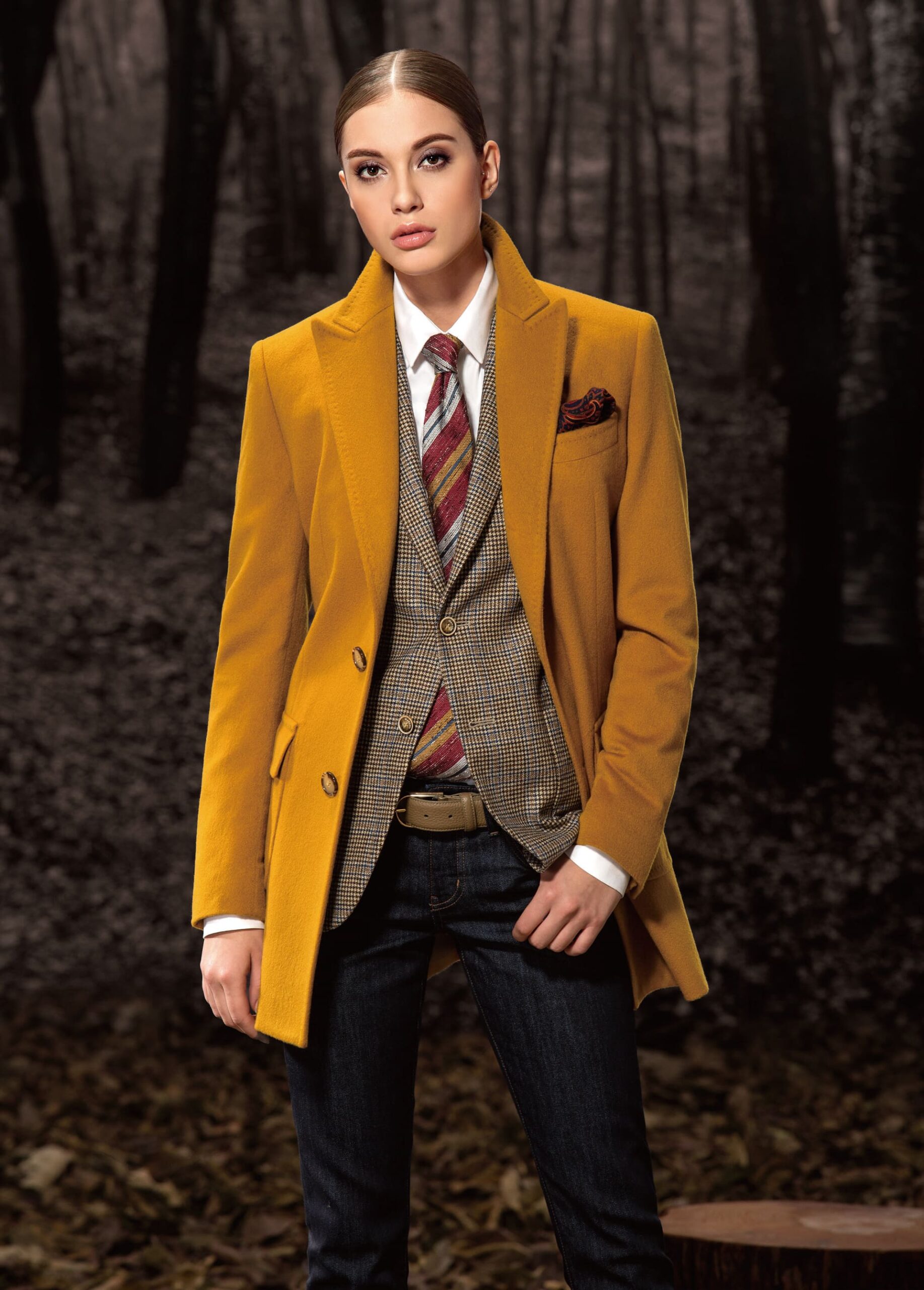 A model wearing a tailored mustard bespoke women’s overcoat, paired with a checkered vest, striped tie, and jeans. The overcoat’s luxurious fabric and precise tailoring create a sophisticated look, perfect for stylish women seeking both warmth and elegance in their wardrobe.